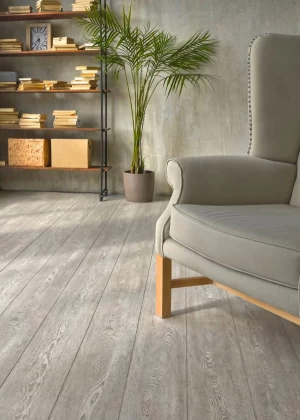 Уютный уголок комнаты с серым креслом, деревянным полом, книжной полкой и большим горшком с пальмой.