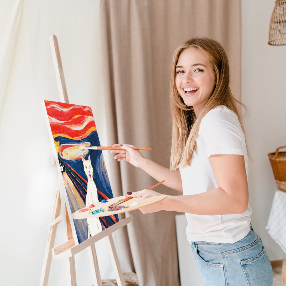 Женщина в белой футболке рисует картину по номерам на холсте, улыбаясь и смотря в камеру, держит палитру с красками в руке.