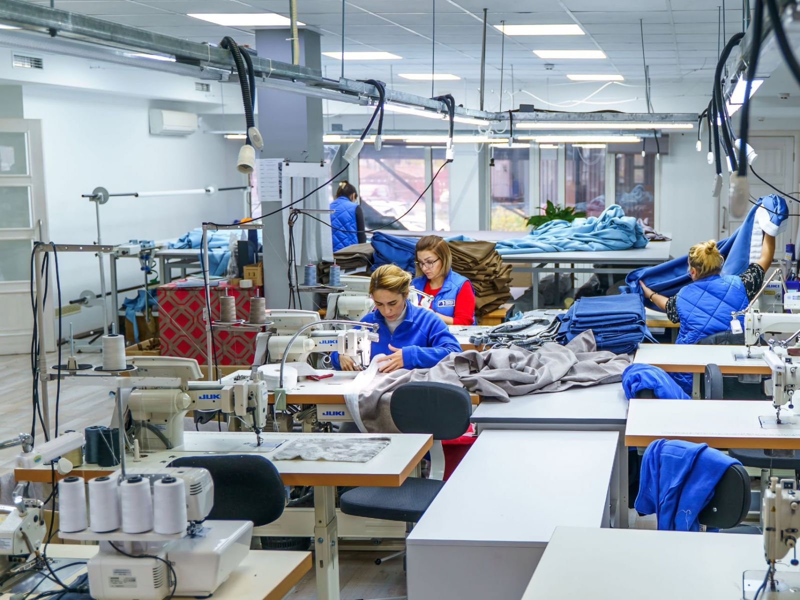Работники на производстве технической ткани в синих халатах занимаются пошивом на промышленных швейных машинах в светлом помещении мастерской