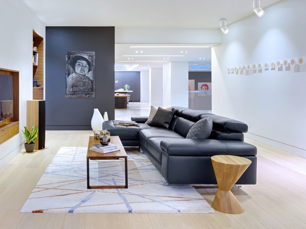 Современная гостиная с черным диваном, деревянным столиком, ковром в голубых тонах. На темной стене висит абстрактное портретное изображение. Освещенное пространство с деревянными элементами и проходами.