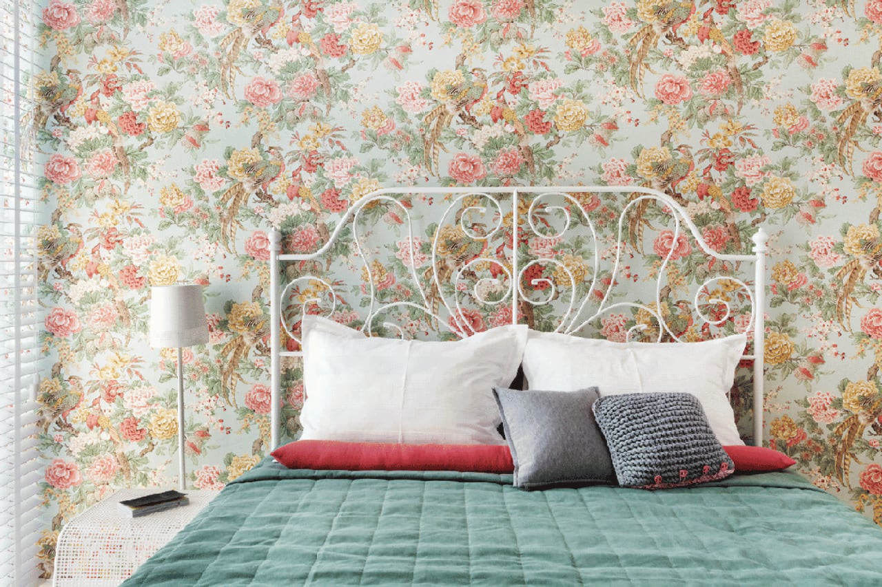 Уютная спальня с кованой белой кроватью. Стена украшена обоями с ярким цветочным узором. На кровати подушки и плед. Рядом стоит настольная лампа и жалюзи.