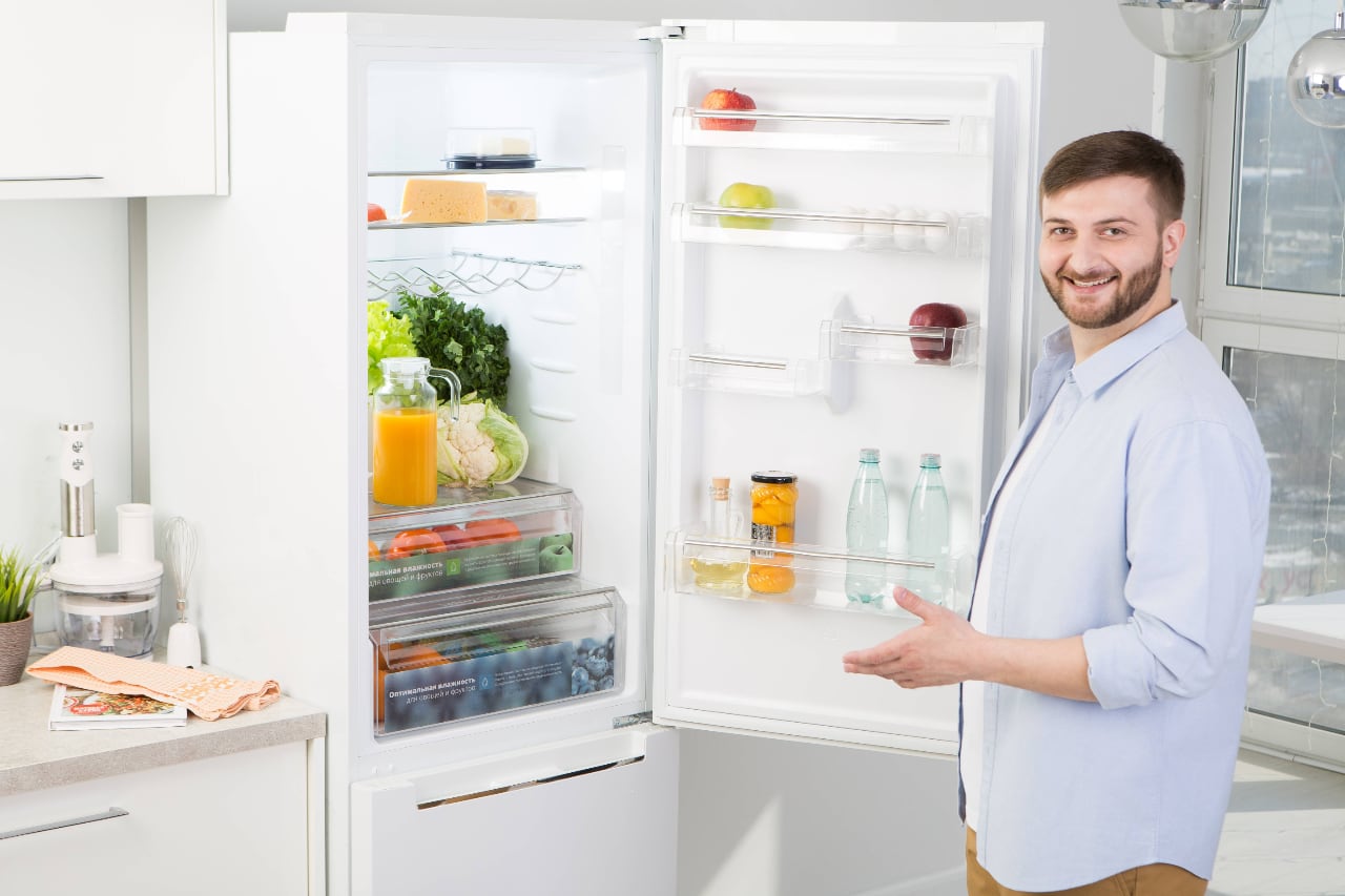Мужчина открывает холодильник, полный свежих продуктов: фрукты, овощи, напитки, сыр, банки, светлая кухня, домашняя атмосфера.