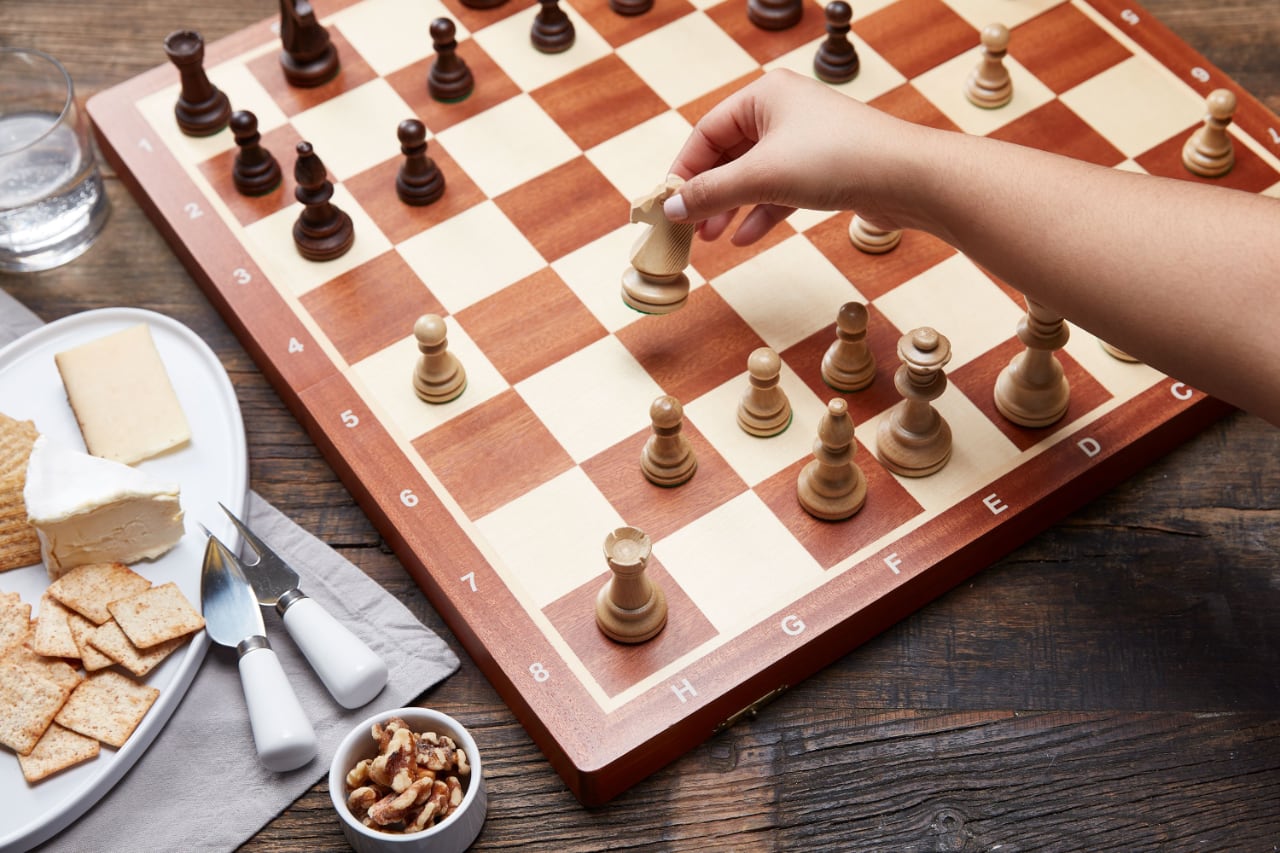 Рука перемещает шахматную фигуру на деревянной доске. Рядом тарелка с сыром, гренками и орехами на старом столе.