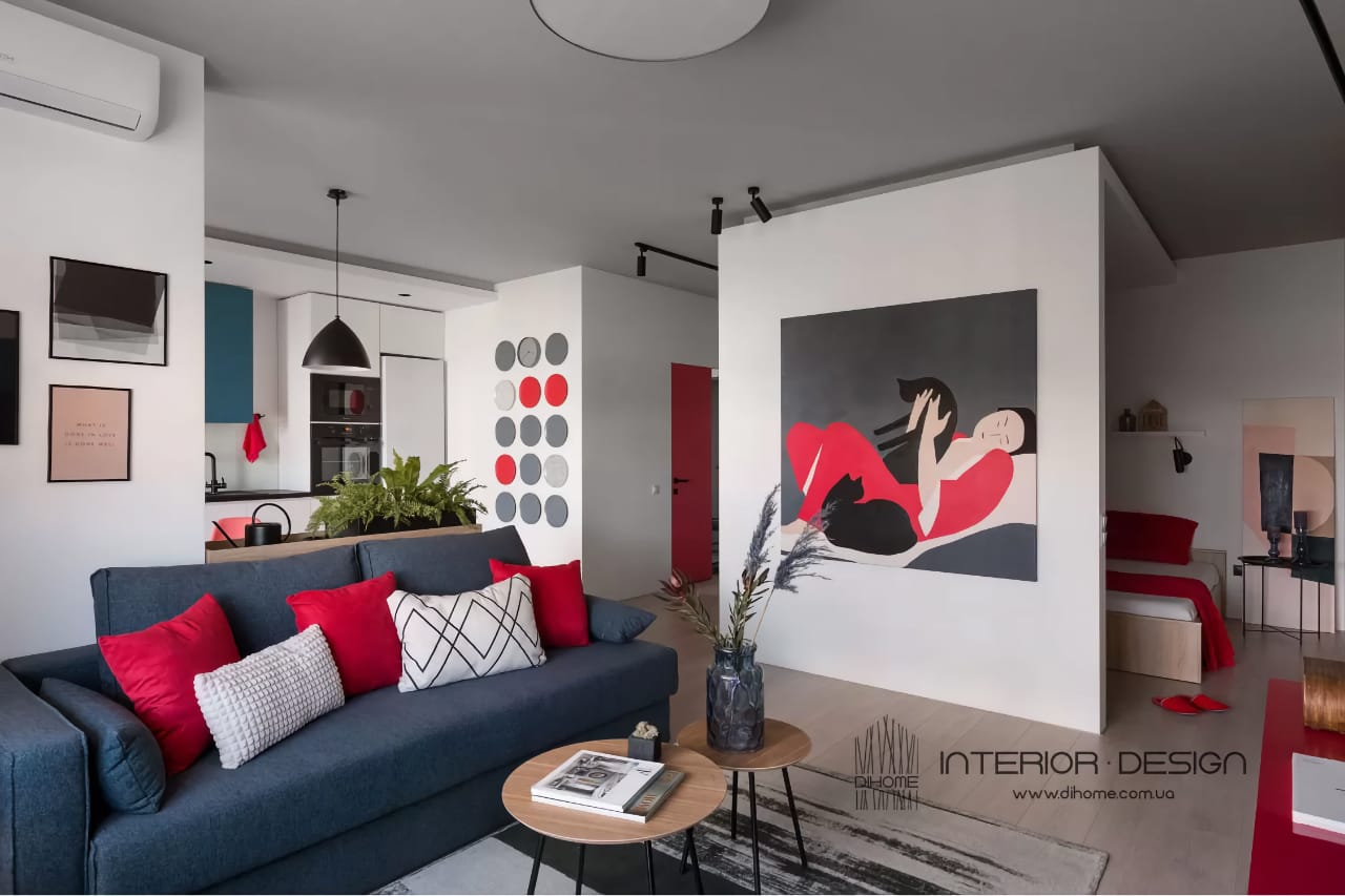 Современный интерьер однокомнатной квартиры. В комнате есть темно-синий диван с красными и белыми подушками, журнальный столик, и большая картина на стене с абстрактным изображением человека в красных брюках. Слева видна кухня с черно-белыми шкафами и красным акцентом. В целом, дизайн комнаты сочетает в себе современные и яркие элементы с акцентом на красный цвет.