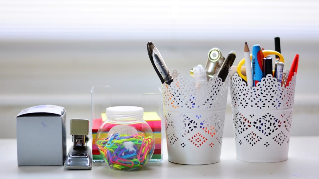 Канцелярские принадлежности: две корзины с ручками, банка с цветными резинками, степлер и скотч на столе.