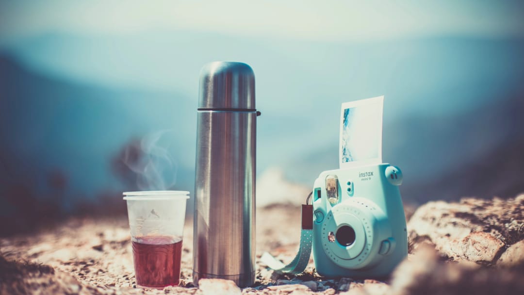 Термос, пластиковый стакан с чаем, мгновенная камера на фоне горного пейзажа с размытым фоном.