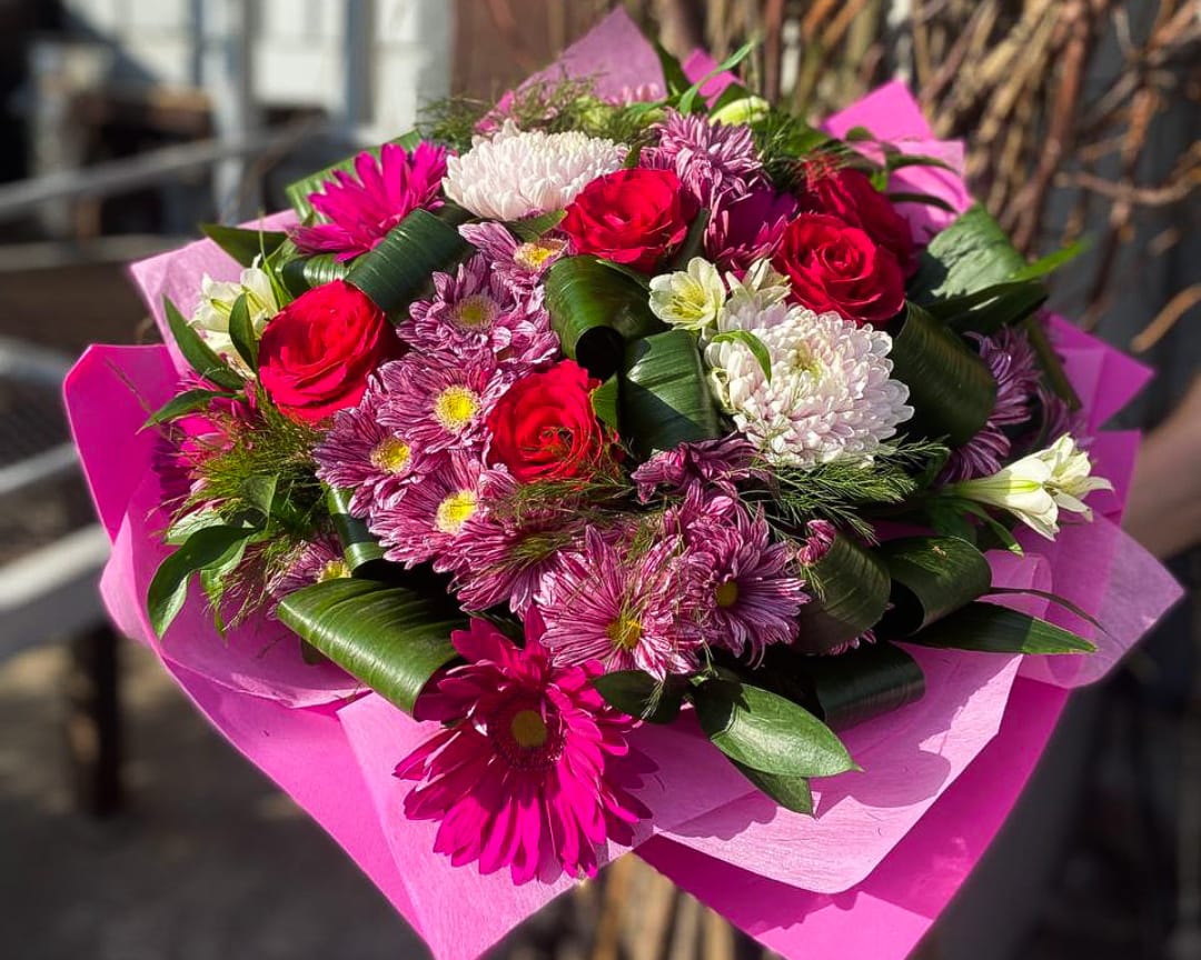 Розкішний букет з червоними трояндами, рожевими хризантемами, герберами та зеленню, упакований у яскраво-рожевий папір, квітковий подарунок на сонячному світлі.