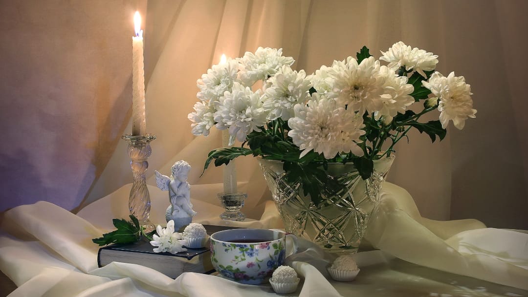 Елегантний стіл з білими хризантемами в кришталевій вазі, свічку, фарфорову чашку і фігурки ангелів на тканинному тлі.