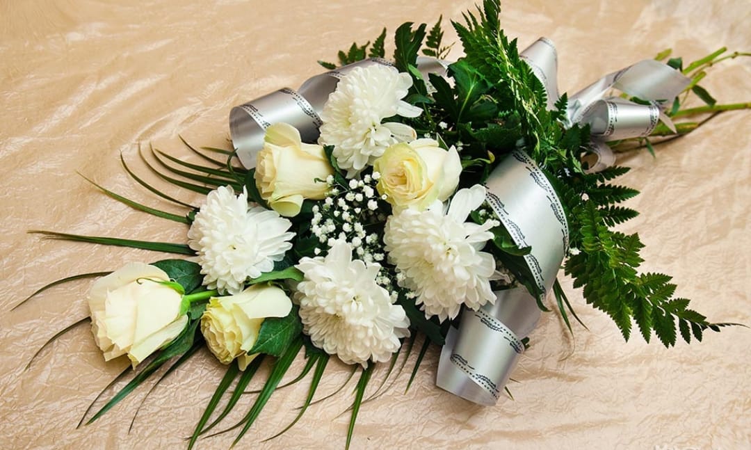 Жалобний букет з білими хризантемами, кремові троянди та зеленню, прикрашений сріблястою стрічкою на бежевому фоні, елегантне квіткове оформлення.