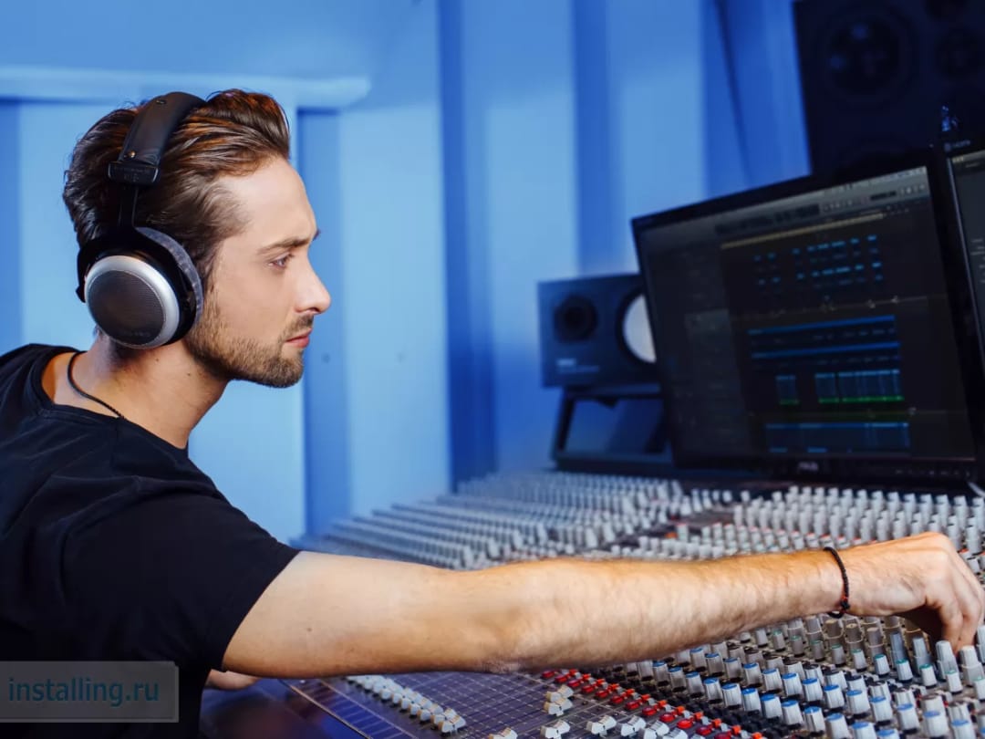 Молодой мужчина в наушниках регулирует оборудование на звуковом микшере в студии с синими стенами и мониторами.