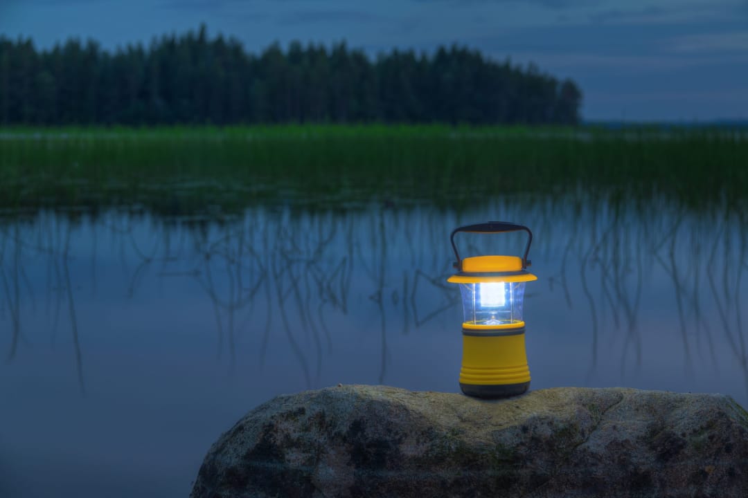 Включенный кемпинговый фонарь на камне возле тихой воды с отражением вечернего леса на фоне закатного неба.