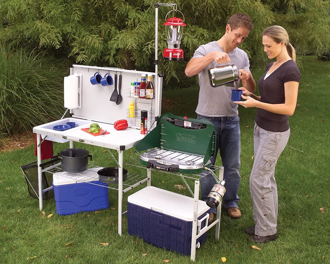 Мужчина и женщина используют мобильную кемпинговую кухню с приборами и посудой на заднем дворе, наливают напиток из термоса.