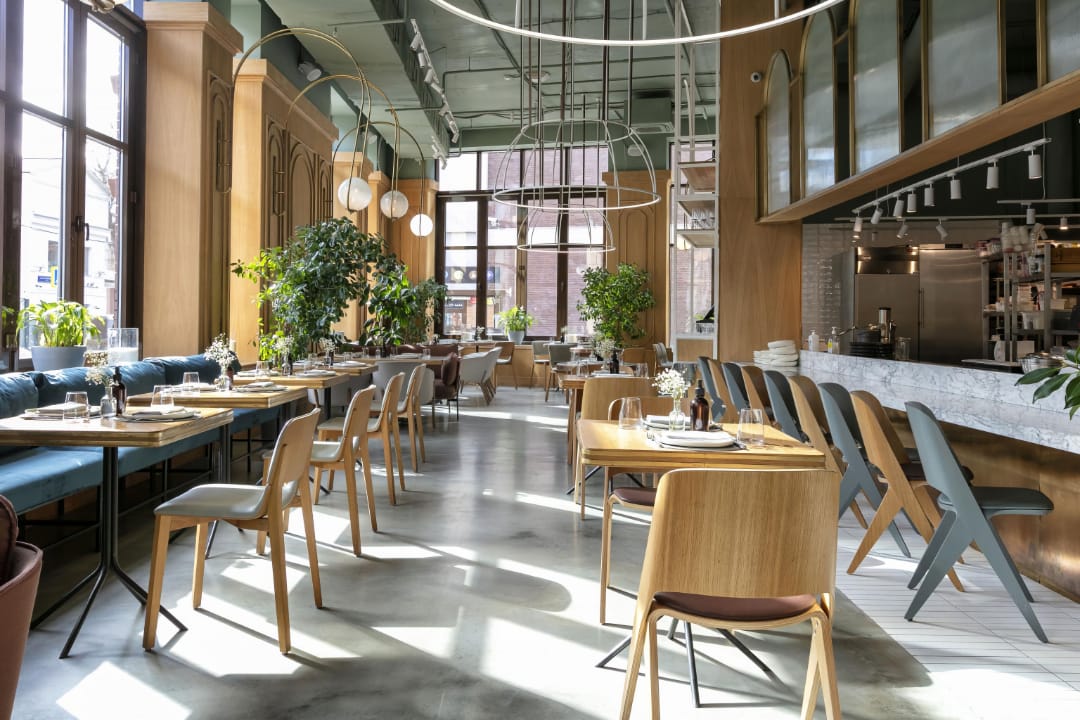 Интерьер просторного кафе с большими окнами, деревянной мебелью, зелеными растениями и стильными подвесными светильниками.