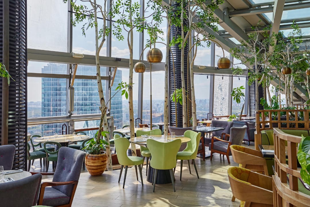 Ресторан с панорамными окнами и видом на город, украшенный живыми растениями, современными мебельными гарнитурами и подвесным освещением.