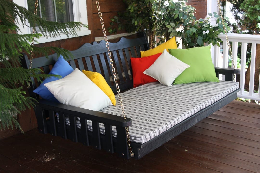 Качели-диван черного цвета с пестрыми подушками на деревянной веранде окружены зелеными растениями и белыми перилами.