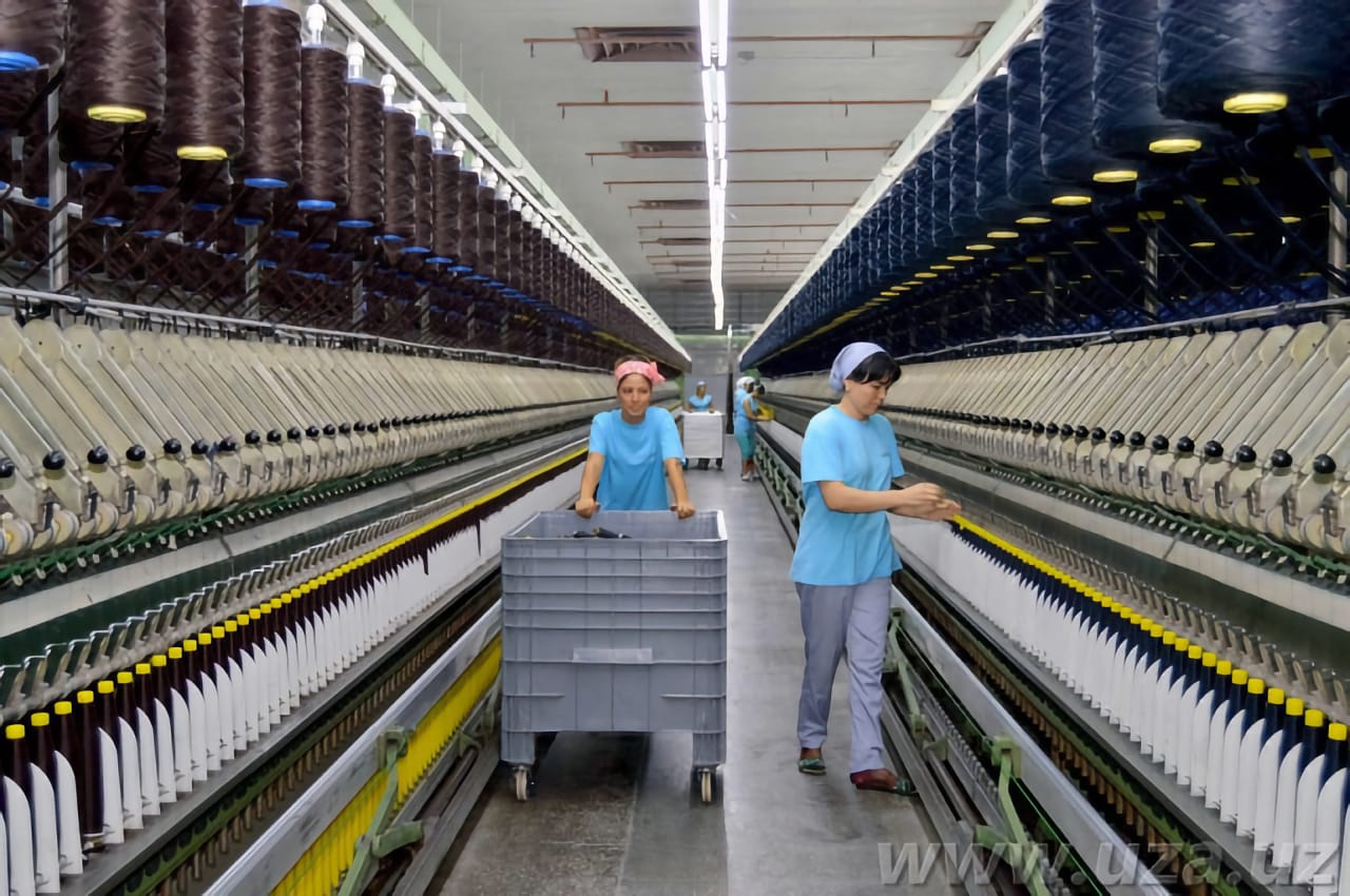 На фотографии изображён текстильный завод, где два рабочих в синей форме обслуживают машины для прядения нитей.
