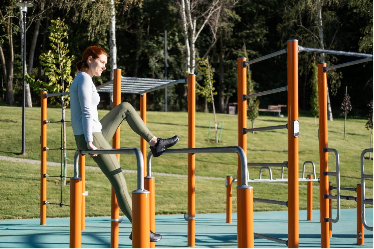Женщина с рыжими волосами в спортивной одежде делает упражнения на уличных тренажерах. Она поднимает ногу, упираясь на металлические перекладины оранжевого цвета. Фон изображения - зеленый газон и деревья в парковой зоне в ясный солнечный день.