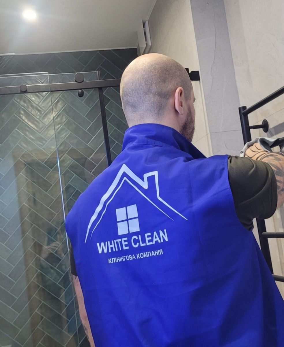 Мужчина в синей униформе с логотипом "WHITE CLEAN" убирает в ванной комнате, отражаясь в зеркальной душевой кабине.