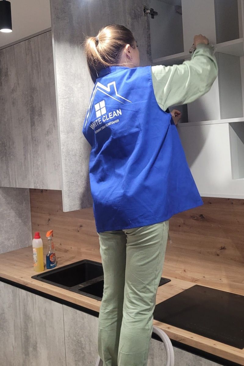 Женщина в синей рабочей униформе с надписью "WHITE CLEAN" убирает на кухне, стоя на стуле.
