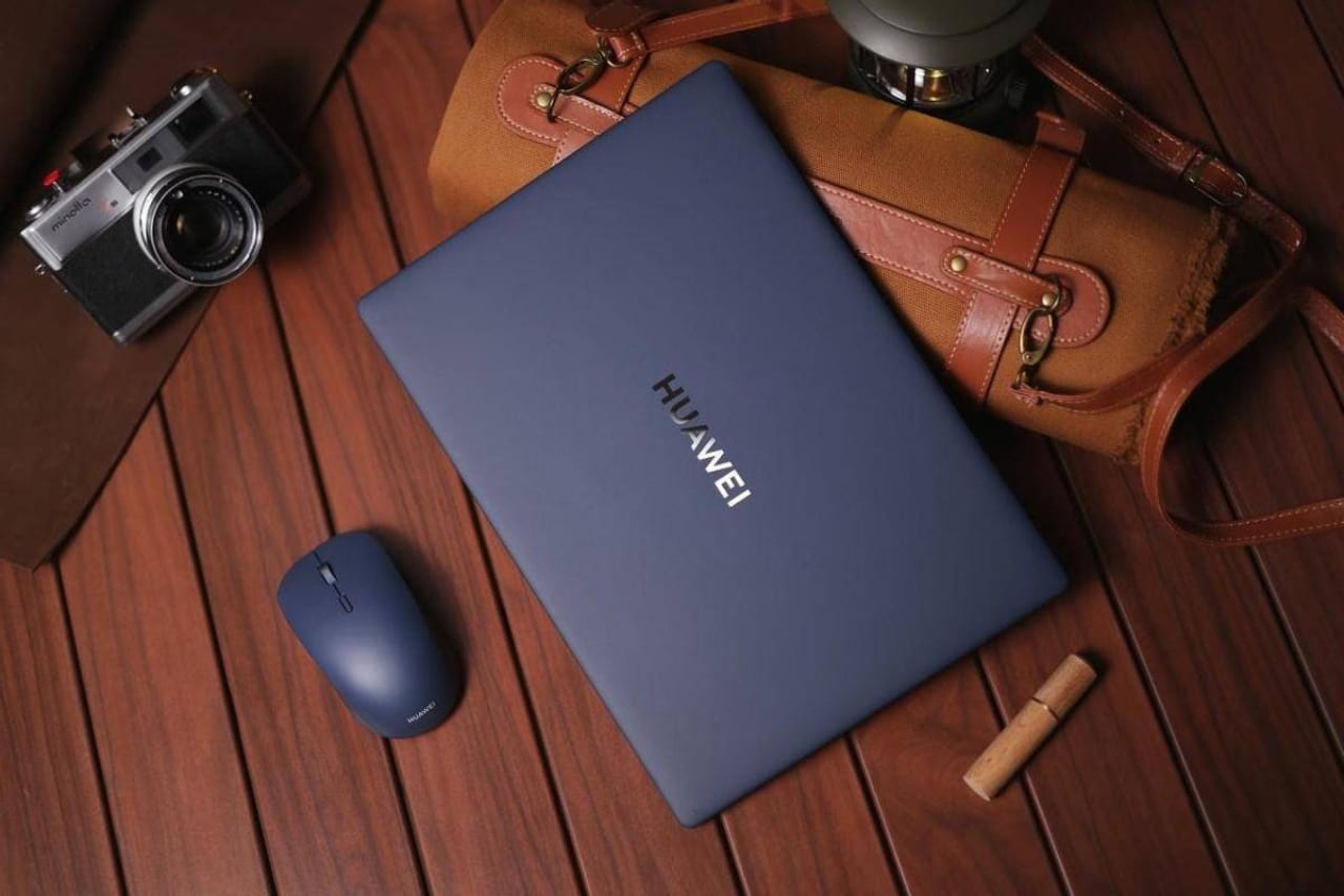 Синий ноутбук HUAWEI, рядом беспроводная мышь, кожаная сумка и старинная камера на деревянном фоне.