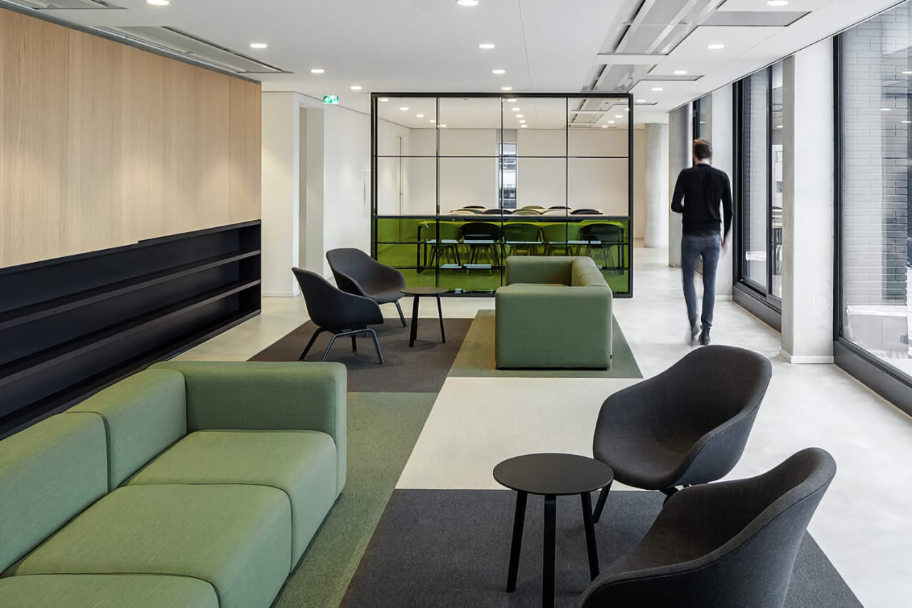 Сучасний офіс із великими вікнами, зеленим диваном, чорними кріслами, людина йде вздовж вікна, зелені акценти.