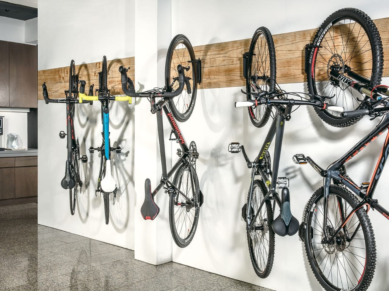 На зображенні представлена стіна усередині приміщення із кріпленнями для велосипедів. На кріпленнях висять чотири велосипеди різного кольору та дизайну. Зліва направо: велосипед із чорною рамою та синіми елементами, велосипед із білою рамою та жовтими елементами, велосипед із чорною рамою та червоними акцентами, та велосипед із чорною рамою та зеленими вставками. Внизу під кожним велосипедом висять відповідні сідла. На задньому плані видно частину кухонного гарнітуру з мікрохвильовою піччю.
