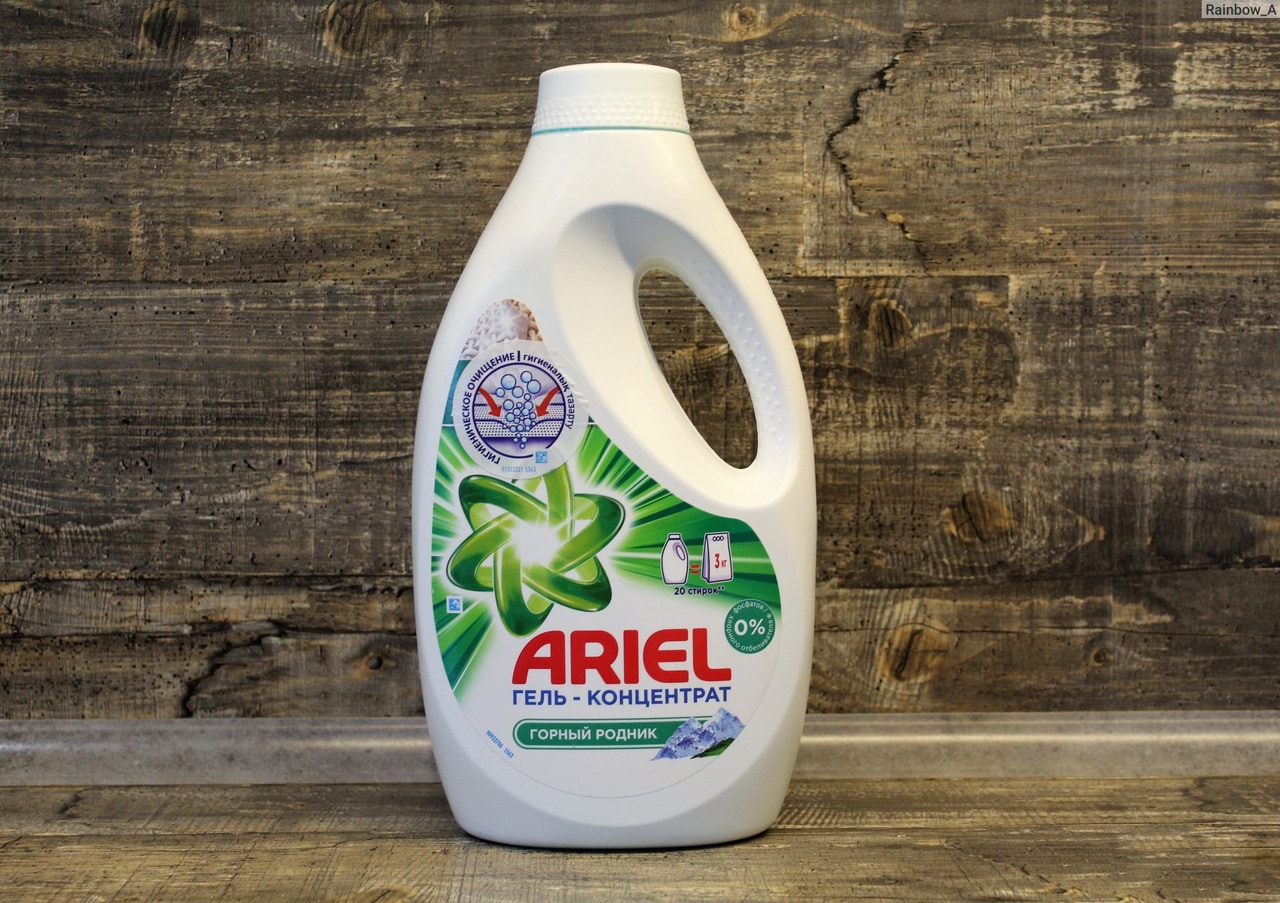 біла пластикова пляшка з миючим засобом марки "ARIEL" із написом "Гель-концентрат" на передній етикетці. Пляшка має унікальну форму з інтегрованою ручкою для зручності використання. Логотип бренду та візуальні елементи, такі як зображення гірського джерела та зелений квітковий мотив, припускають свіжість та ефективність продукту. Етикетка також вказує, що у складі немає фосфатів (0% фосфатів), і є маркування, яке, ймовірно, вказує на обсяг або кількість прань. Фон за пляшкою - текстуроване дерево, що додає зображення екологічний та натуральний контекст.
