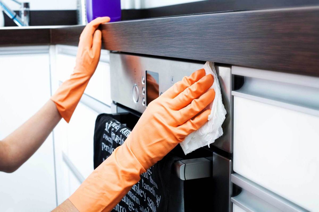 Лицо в оранжевых резиновых перчатках протирает белую салфетку нержавеющую поверхность микроволновки, установленную между шкафами на кухне.