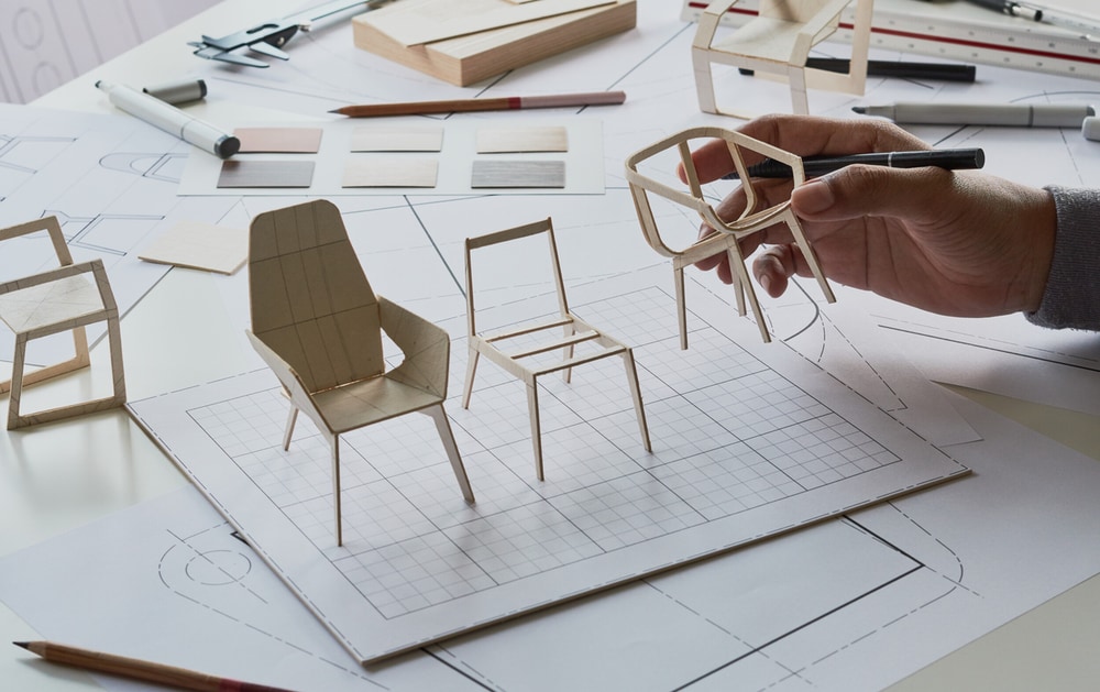 Рабочий стол, который смотрится как рабочее место дизайнера мебели либо конструктора. На столе разбросаны чертежи, среди которых виден план кресла, и есть несколько макетов стульев, сделанных из картона или древесины. Рука держит один из макетов стульев, а на столе есть инструменты для чертежа, такие как линейки, карандаши и что-то, что выглядит как резцы или инструменты для обработки макетов. Весь этот сценарий передает впечатление процессу проектирования мебели.