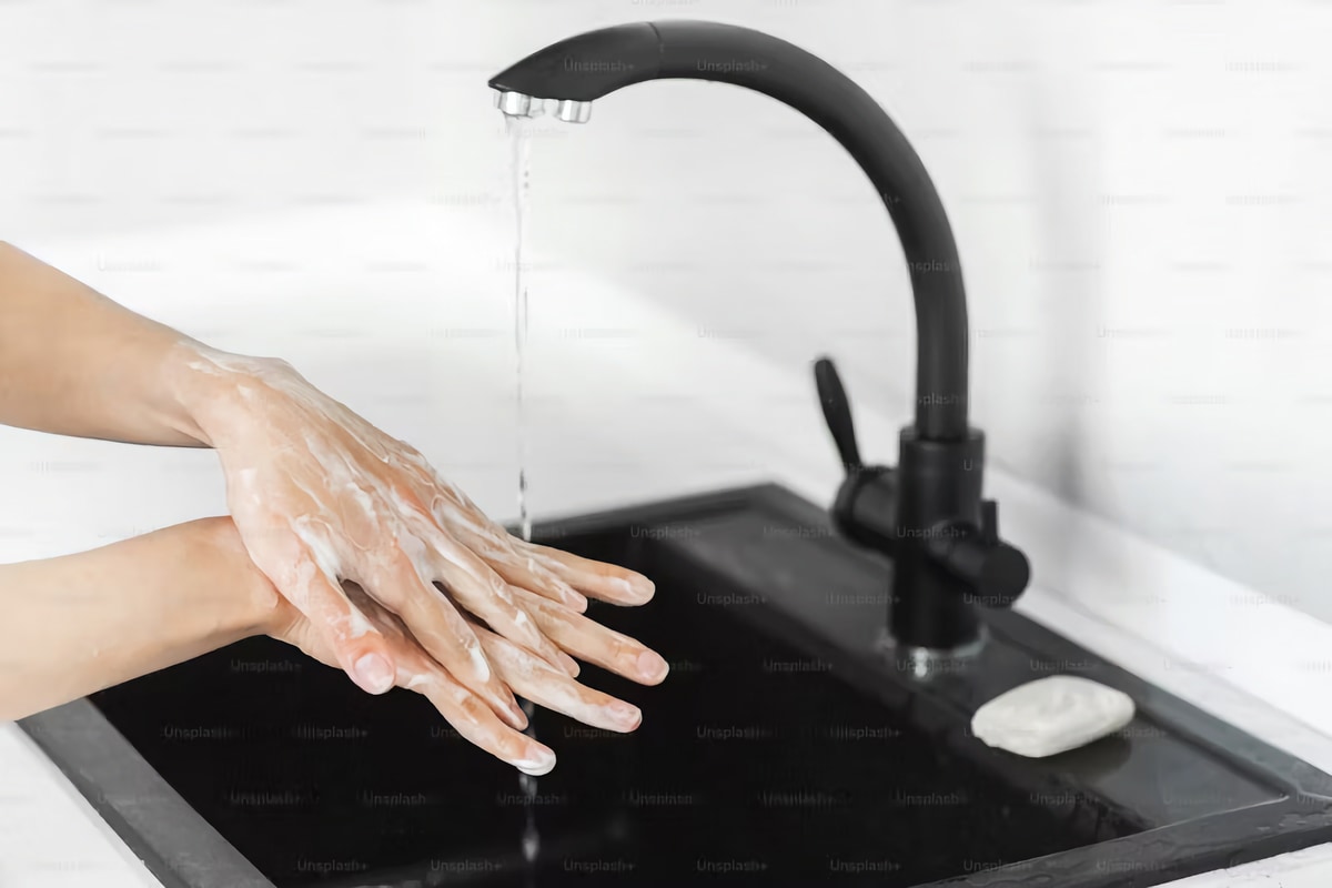 Руки людини, які миє під струменем води з чорного сучасного водопровідного крана. Кран встановлений у чорну кухонну раковину. У правому нижньому кутку видно шматочок мила білого кольору. Фон складається з білої стіни з горизонтальною фактурою. Сцена освітлена, створюючи акцент на дії миття рук та гігієни.