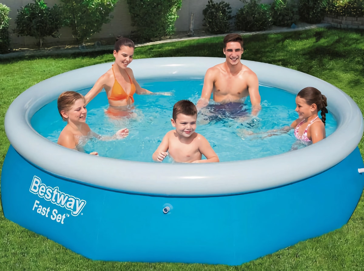 Семья весело проводит время в надувном бассейне синего цвета в саду. Двое взрослых и трое детей улыбаются и играют в воде, окружённые зелёной травой и растениями в солнечный день.