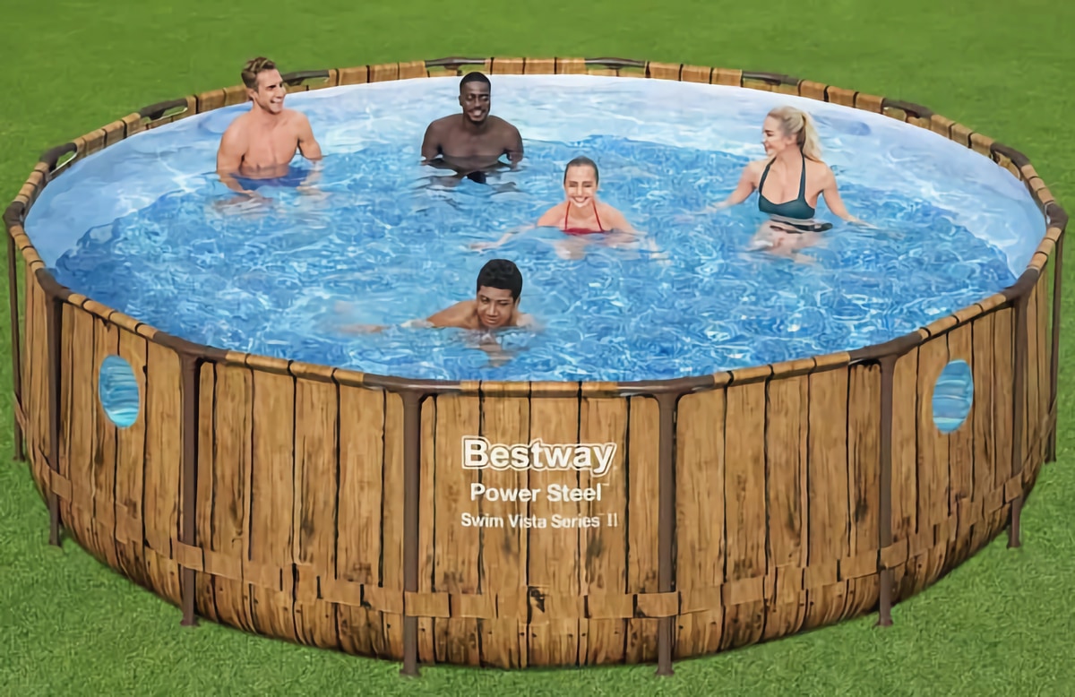 Надувной бассейн, имитирующий деревянное обрамление, установленный на зеленом газоне. В бассейне с водой синего цвета находятся пять человек, которые, по всей видимости, наслаждаются купанием.