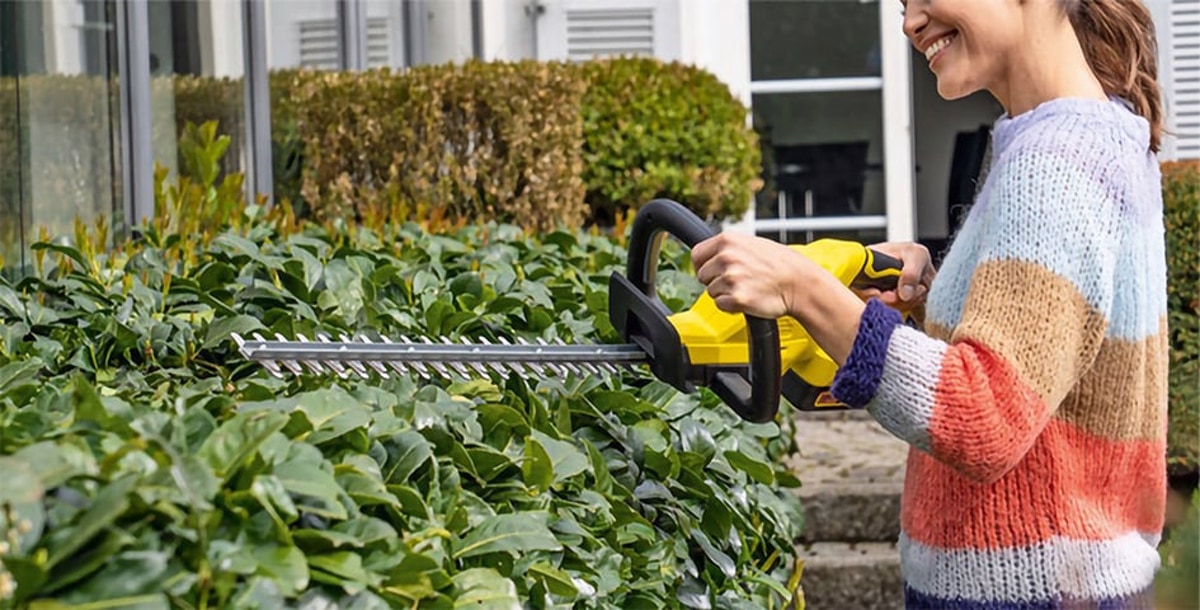 Женщина в цветном свитере стрижет живую изгородь в саду с помощью желтого электрического садового триммера. На фоне видна часть дома и другие зеленые растения. Сцена выглядит дружелюбной и спокойной, иллюстрируя бытовые садовые работы.