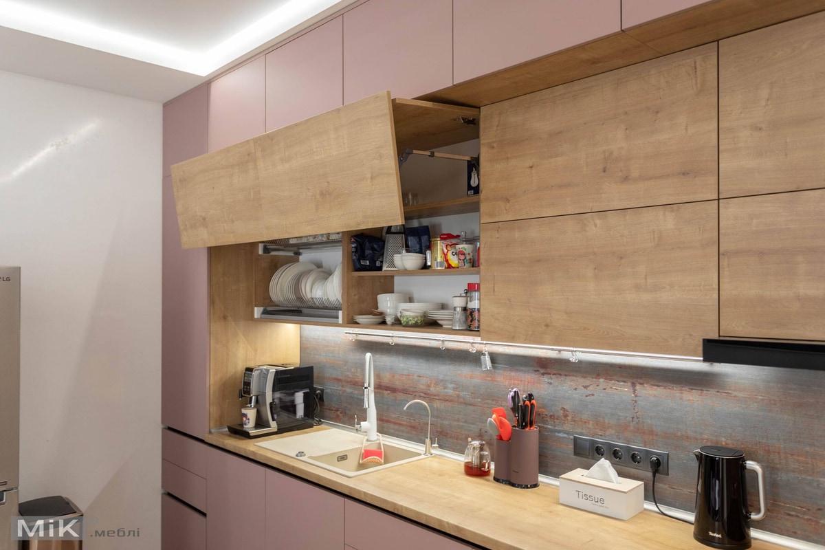 Сучасний кухонний інтер'єр зі світло-рожевими нижніми шафами та дерев'яними верхніми шафами, одна з яких відкрита, щоб показати посуд і приладдя. Тут є кавоварка, раковина з високим краном, підставка для посуду з різними кухонними інструментами, коробка з написом "Серветки" та чорний електричний чайник. Задній фон має рустикальне металеве покриття. Дизайн вирізняється чіткими лініями, а підсвічування під шафою додає простору теплого сяйва.