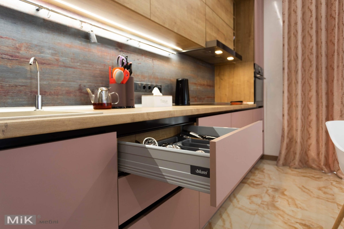 Частина кухні з кремовим мармуровим підлогою та рожевими фасадами меблів. На передньому плані відкритий висувний ящик з кухонним начинням і системою для зберігання. Кухонний стільник з дерев'яним візерунком, на якому розміщені прозорий чайник і стаканчики, чорний електричний чайник та стенд з кухонними приладами оранжевого кольору. Задній фон прикрашає дерев'яна стінка з текстурою та вбудоване світло під верхніми шафами. Зліва помітна частина рожевої занавіски.