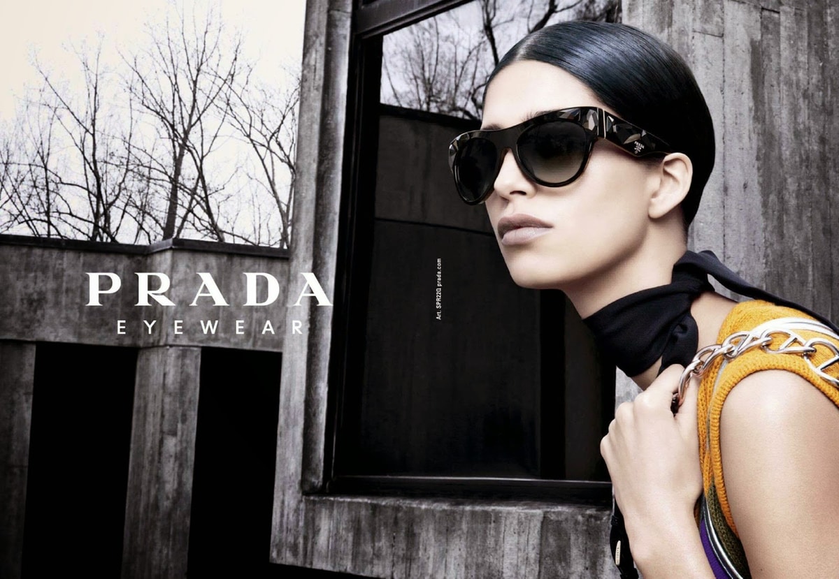 Жінка з темним волоссям і великими сонцезахисними окулярами, спершись на темну будівлю, на задньому плані дерева без листя; напис "PRADA eyewear" у лівій частині зображення.