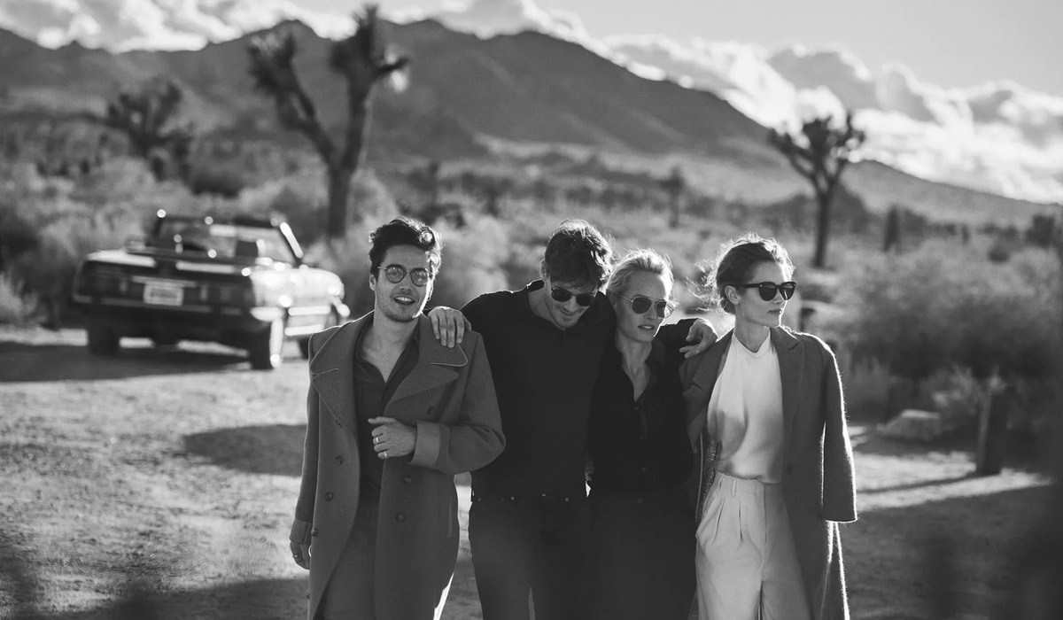 Четыре человека в солнцезащитных очках идут в объятиях на фоне пустынного ландшафта и припаркованного автомобиля, черно-белое изображение.