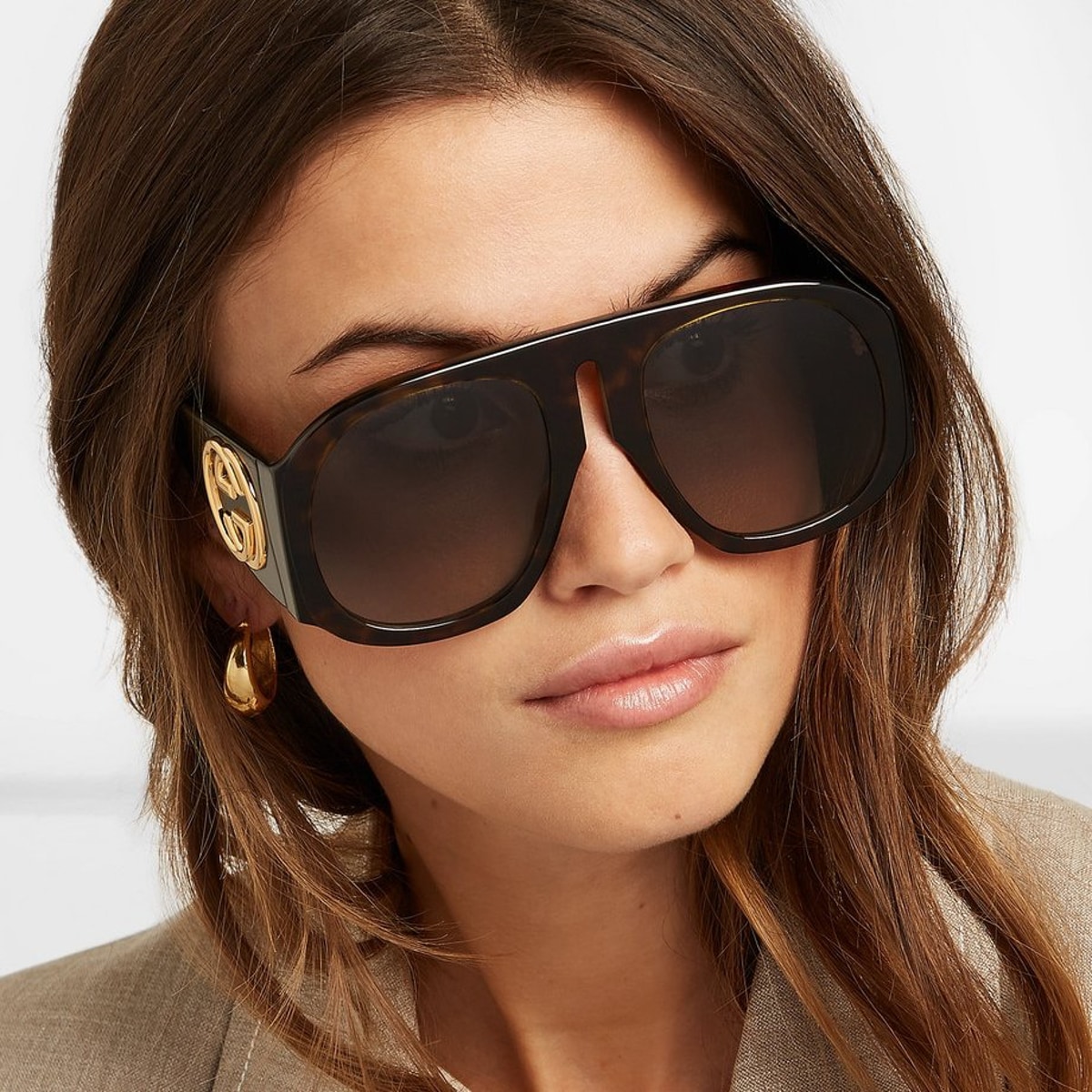 Женщина в больших квадратных солнцезащитных очках с золотым логотипом на дужке, в сером пиджаке, видна половина лица, крупный план.