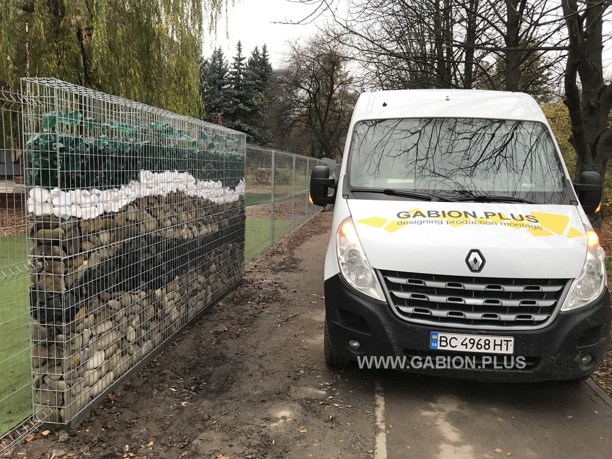 Паркан з габіонів, заповнений камінням і різнокольоровим склом, поруч із службовим автомобілем